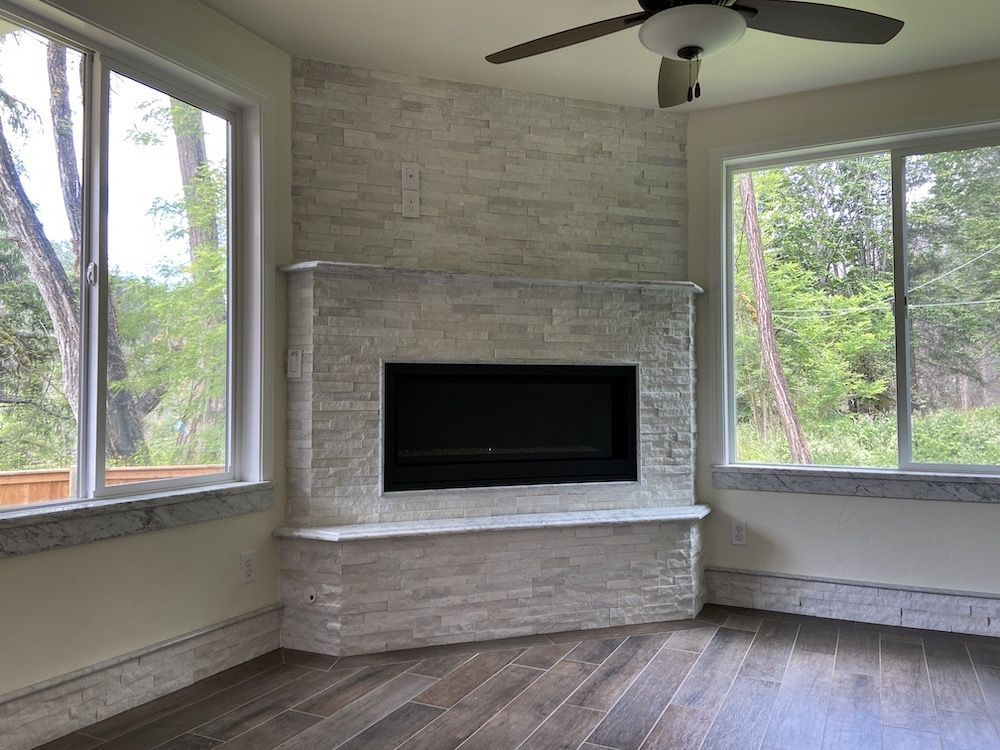 Room with a stone fireplace, windows, ceiling fan, and hardwood floors.