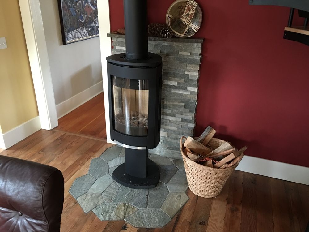 Modern wood-burning stove with stone surround and wood basket on wood floor next to red wall.