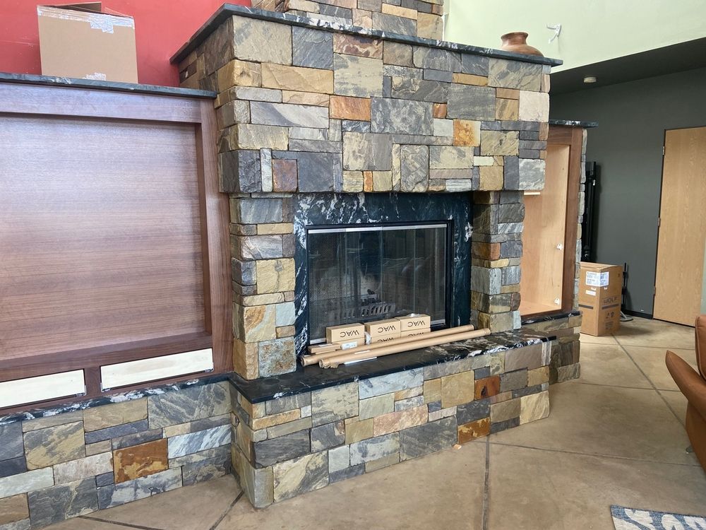 Stone fireplace under construction in a room, with brown stone and black firebox.