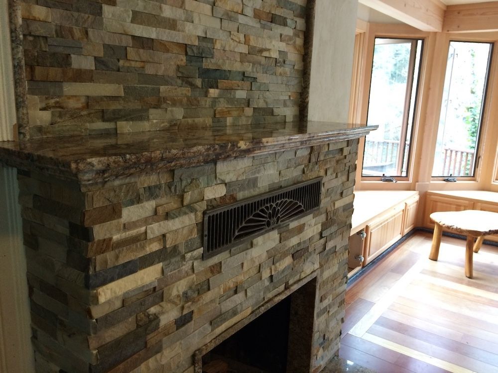 Stone fireplace with a granite mantel, next to windows with a sitting area and a wooden table.