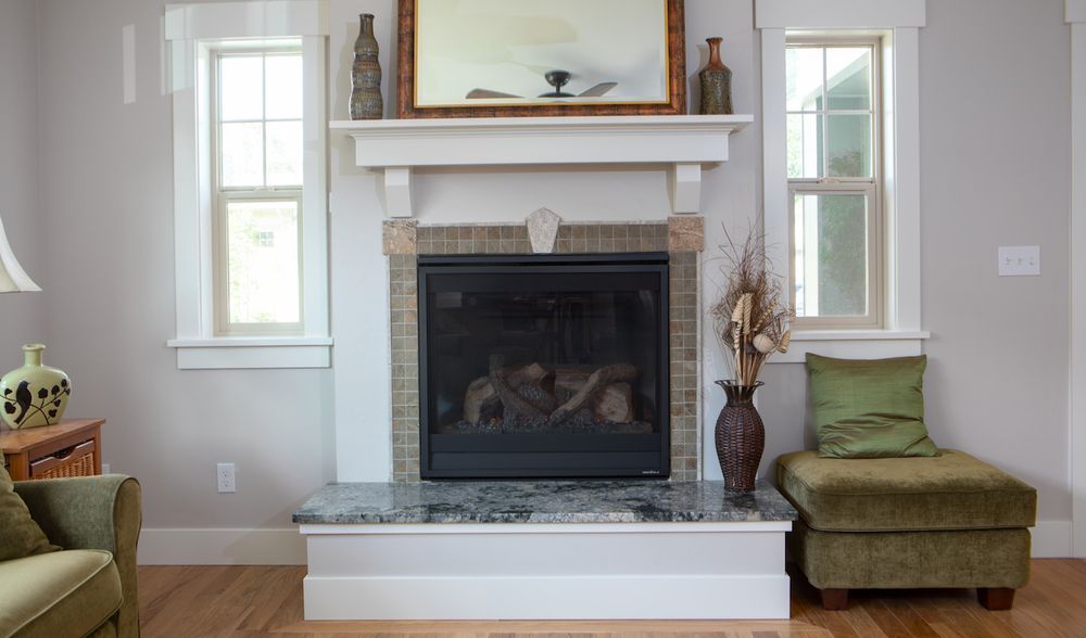 Fireplace flanked by windows, with a mantle and granite hearth, near a green ottoman.