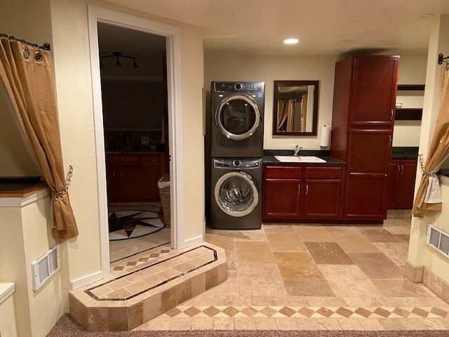 Laundry room with stacked washer/dryer, sink, cabinets, and a doorway. Beige and brown tones.