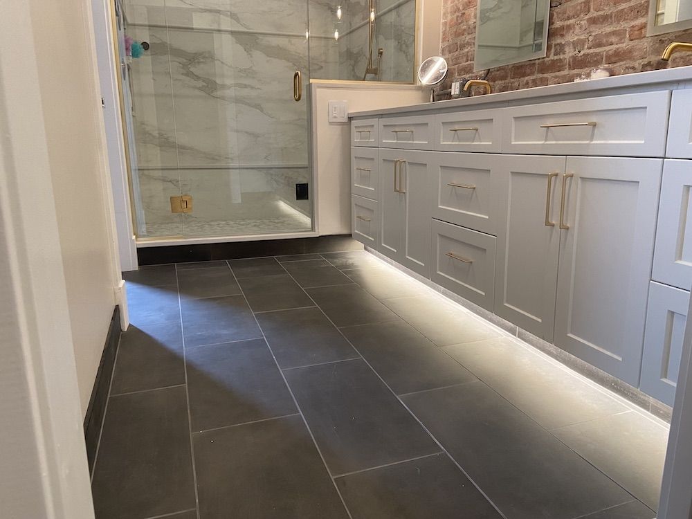 Bathroom with gray tile floor, white cabinets with under-cabinet lighting, glass shower, and exposed brick wall.