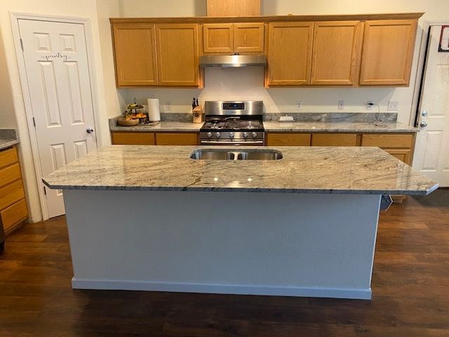 Kitchen with granite island, cabinets, stove, and sink. Wood cabinets and floors with white island base.