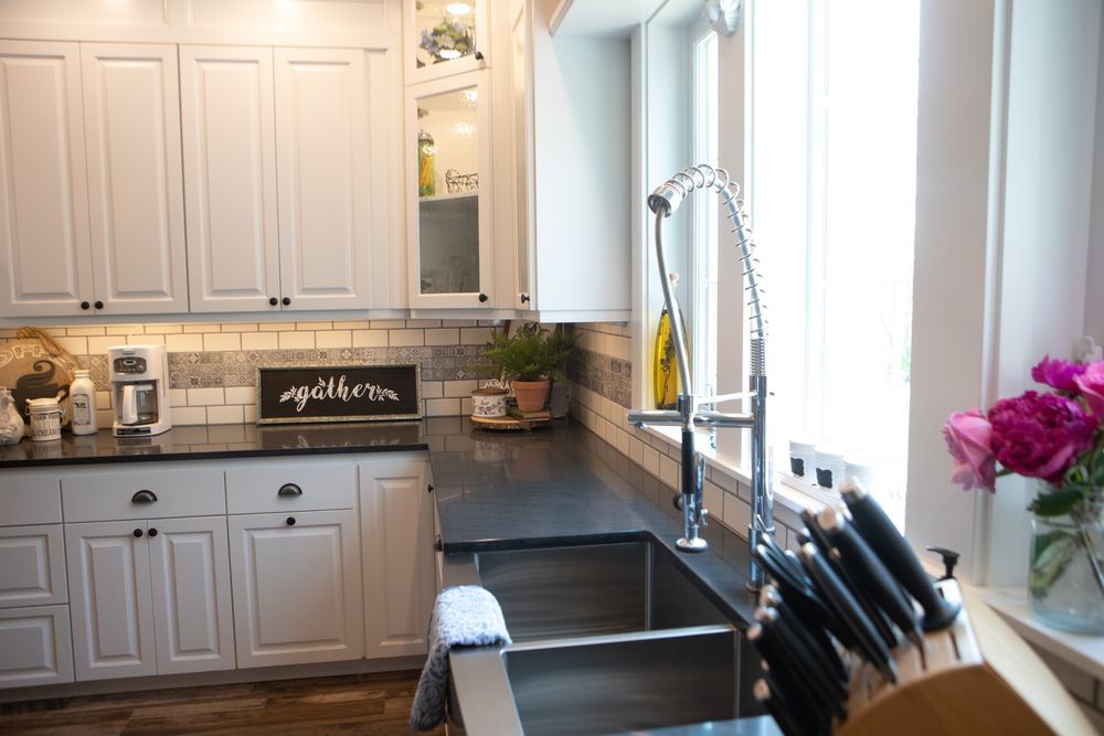 White kitchen cabinets and dark countertops with a stainless steel sink, near a window with sunlight.