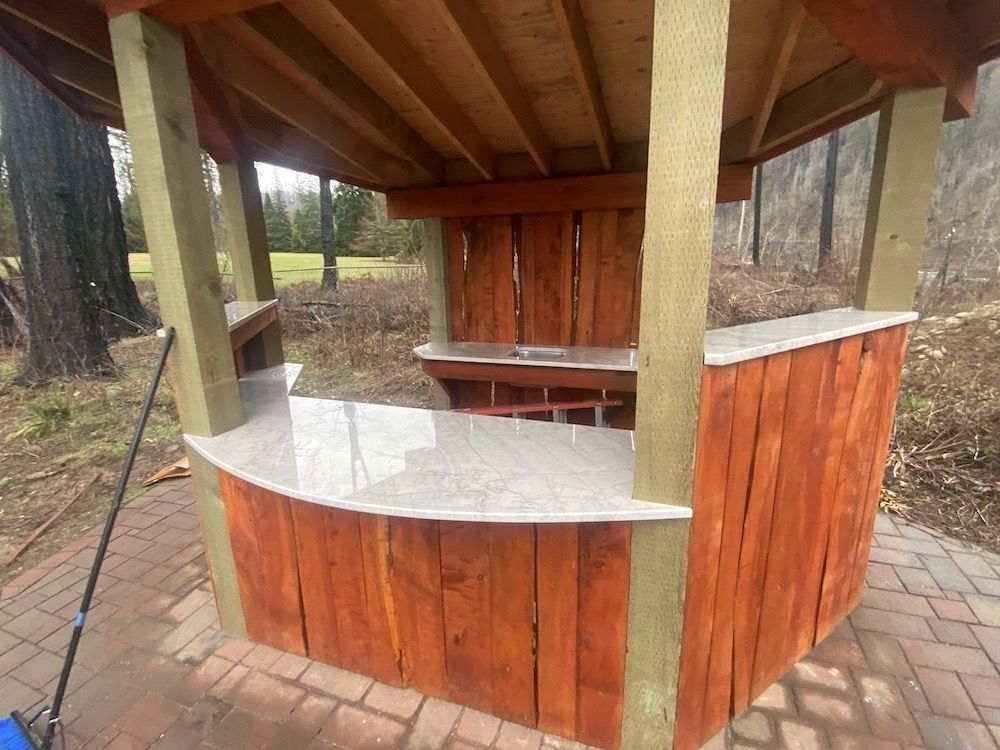 Wooden outdoor bar with granite countertop, set in a gazebo, on a brick patio.