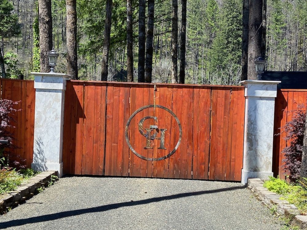 Wooden gate with metal logo, flanked by stone pillars and a gravel driveway.