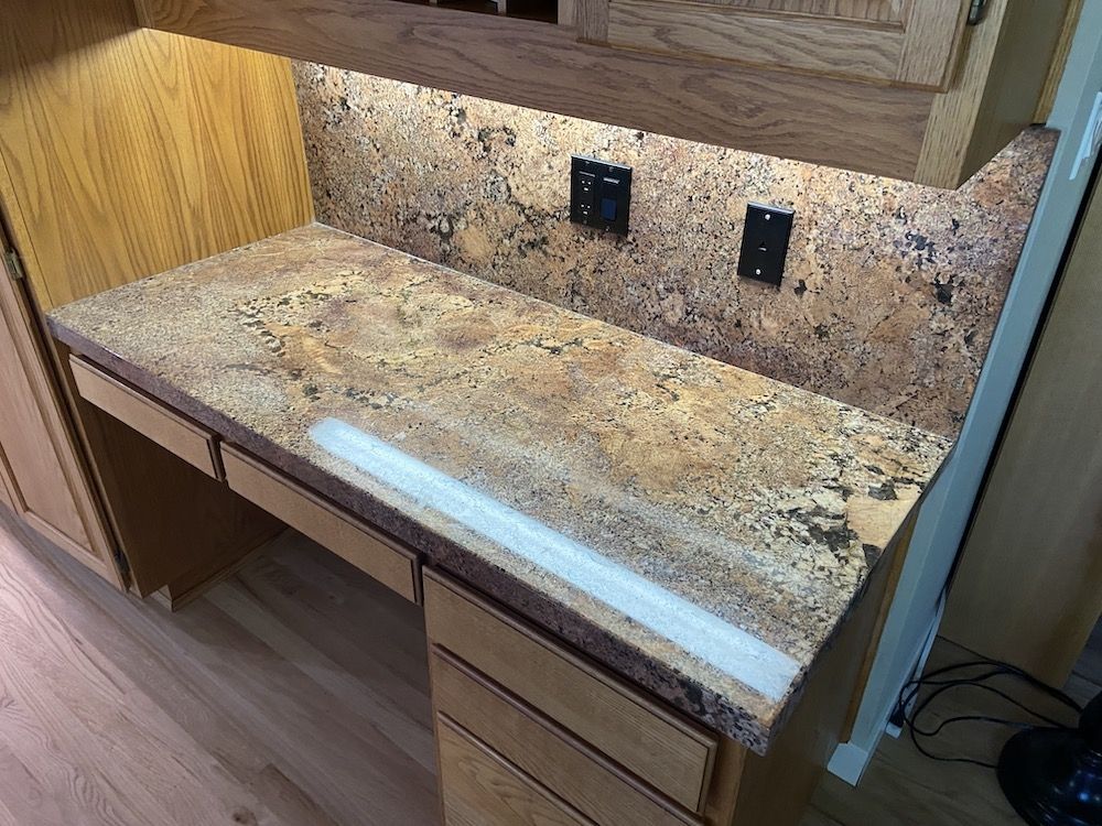 Desk with granite countertop, built-in drawers, and backsplash with power outlets. Overhead cabinet and under cabinet lighting.