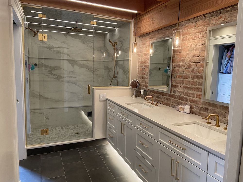 Modern bathroom with white cabinets, brick wall, glass shower, and gold fixtures.