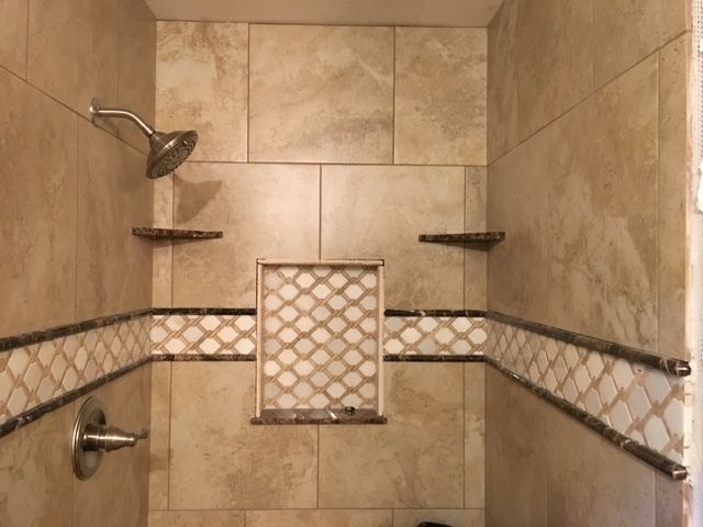 Tiled shower with shower head, shelves, and decorative mosaic trim. Beige and brown tones.