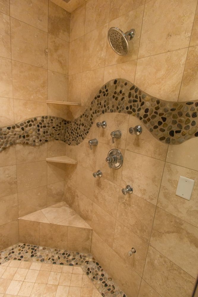 Beige tiled shower with pebble stone accents and multiple shower heads and built-in shelves.