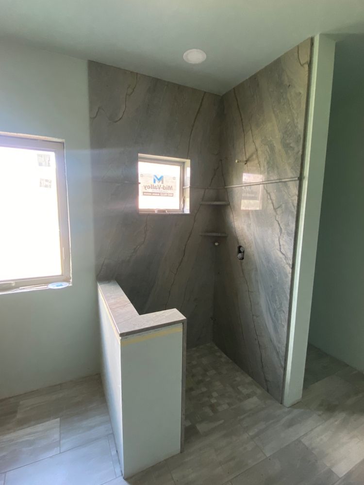 Unfinished bathroom with gray tiled shower, window, and small wall; light-colored walls and floor.