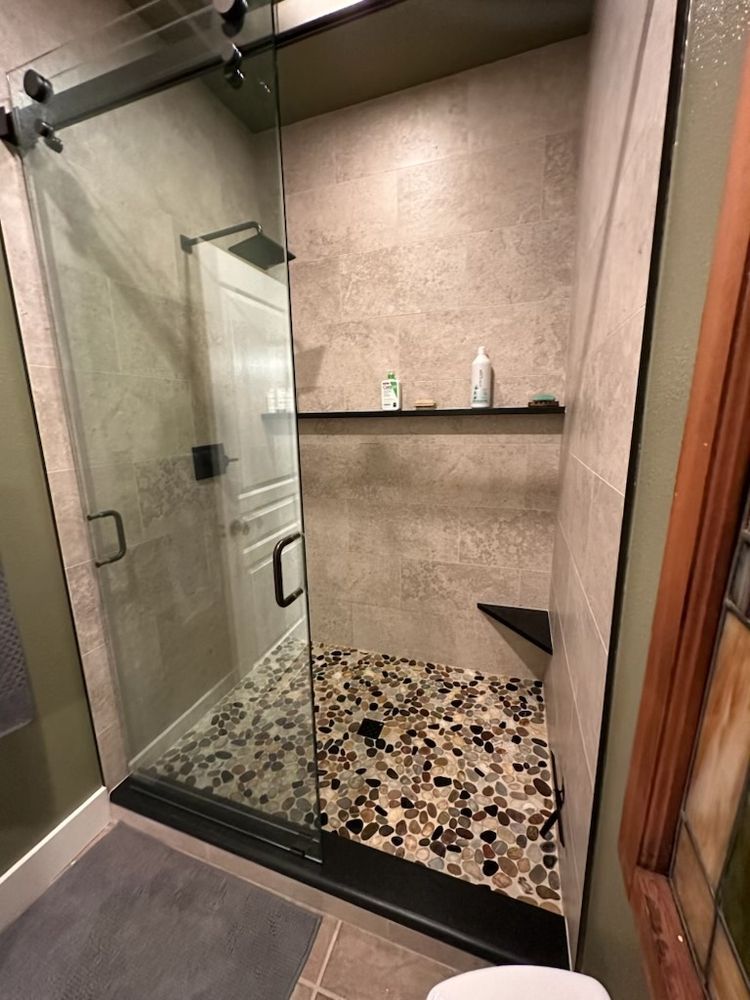 Shower stall with pebble floor, glass door, and built-in shelf; gray and brown tones.