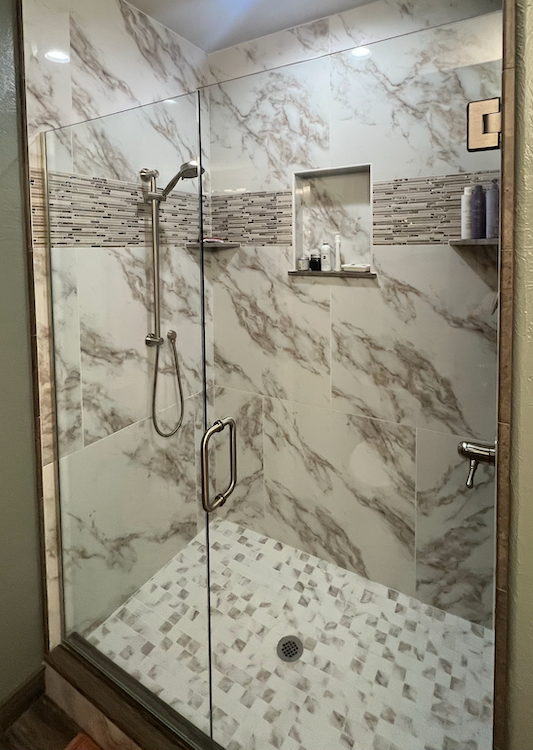 Glass-doored shower with marble-patterned tile, stone floor, and recessed shelf.