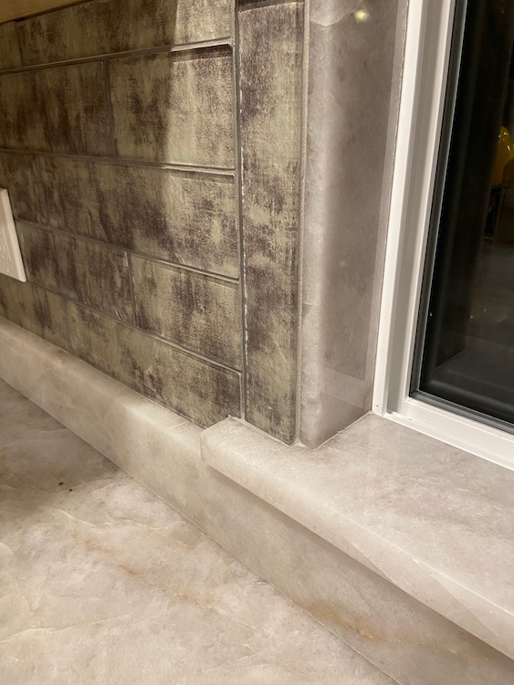 Close-up of a wall with textured tiles and marble trim near a window.