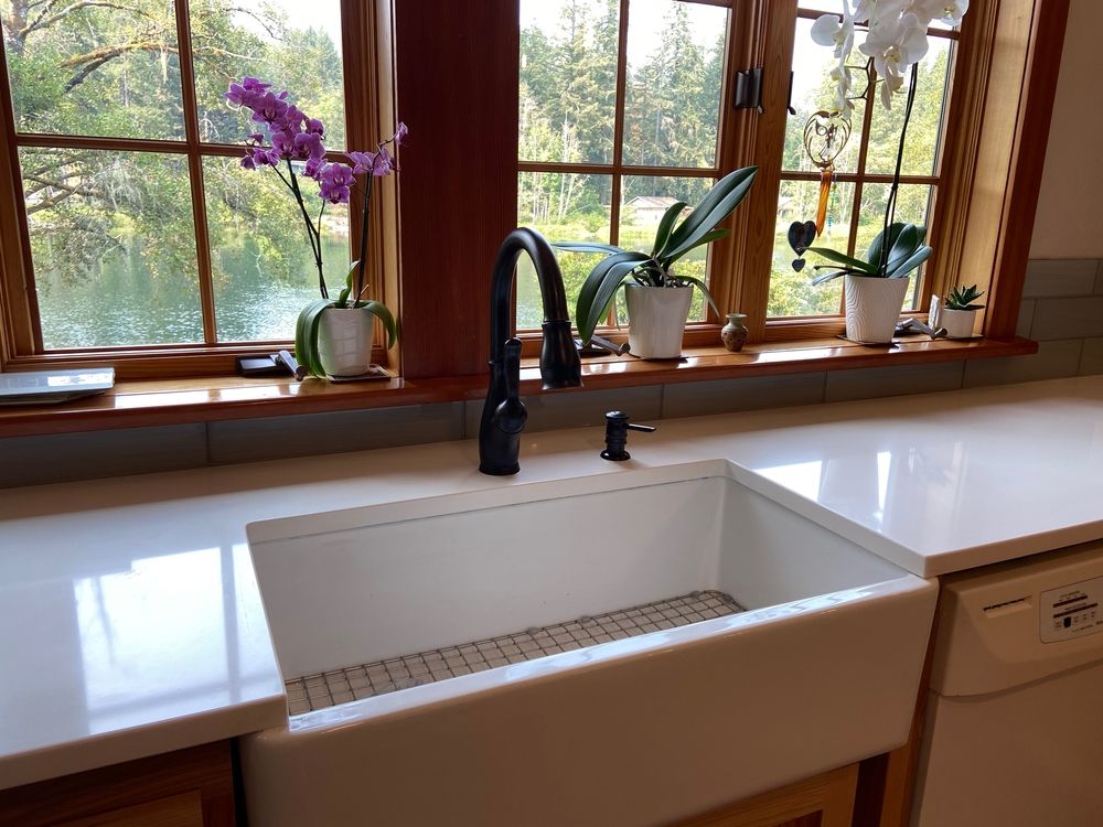 Kitchen sink with view of trees, orchids on windowsill, black faucet, white countertop.