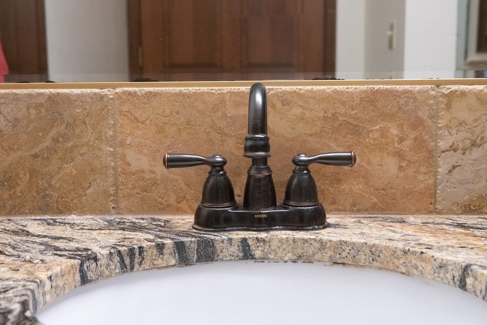 Close-up of a dark faucet with two handles on a speckled countertop and tiled wall, near a white sink.