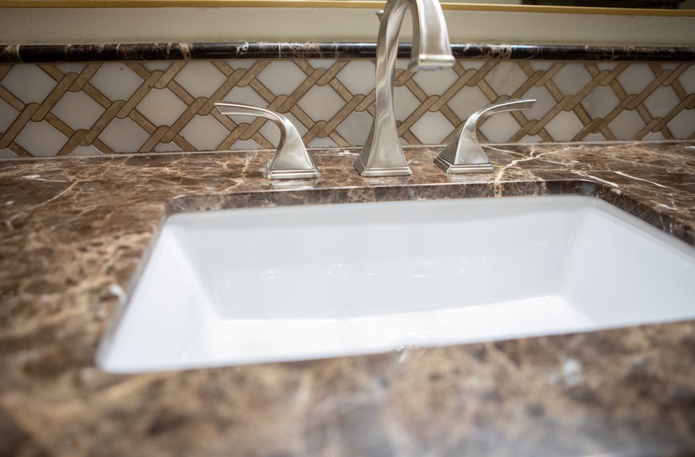 White sink in a bathroom, set in a brown marbled countertop with silver faucet and decorative backsplash.