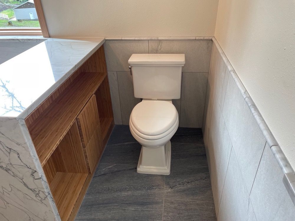 Toilet in a bathroom alcove with gray tile floor, light beige walls, and wood-toned cabinets.
