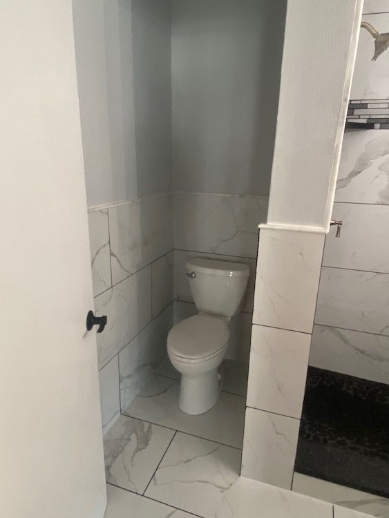 Toilet in a small alcove with white marble-like tile walls and floor. Black door and shower visible.