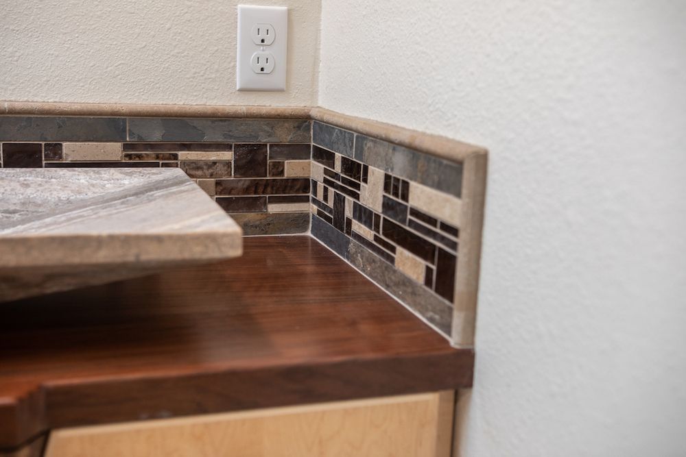 Corner of a bathroom counter with brown and beige tiles and a wooden cabinet. A wall outlet is visible.