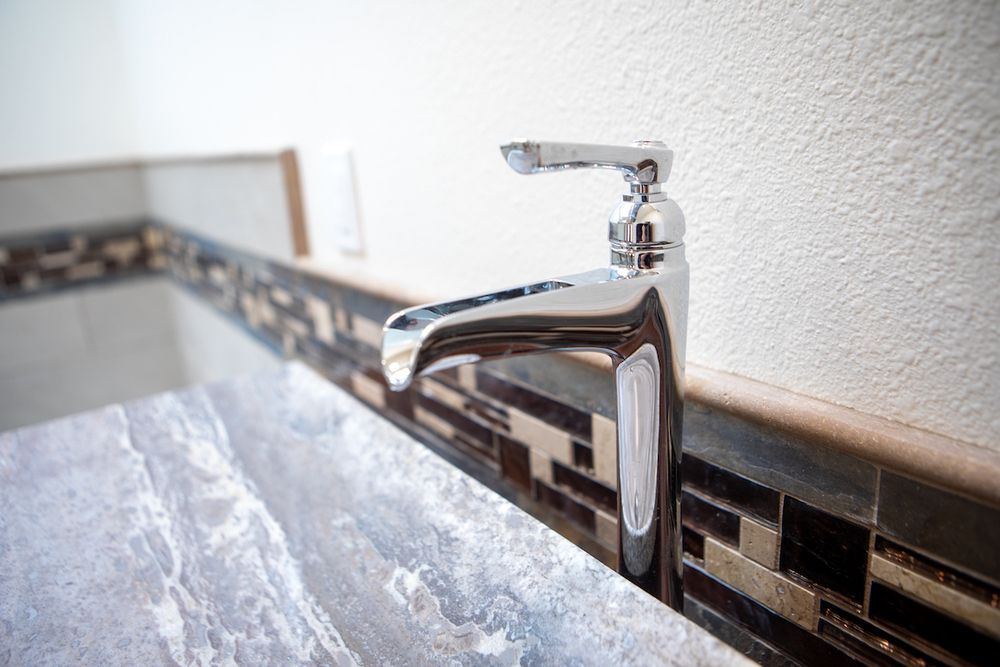 Close-up of a shiny chrome faucet on a marble-like countertop, with a decorative tile backsplash.