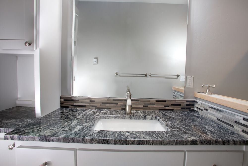 Bathroom vanity with granite countertop, square sink, and large mirror. White cabinets and a mosaic backsplash.