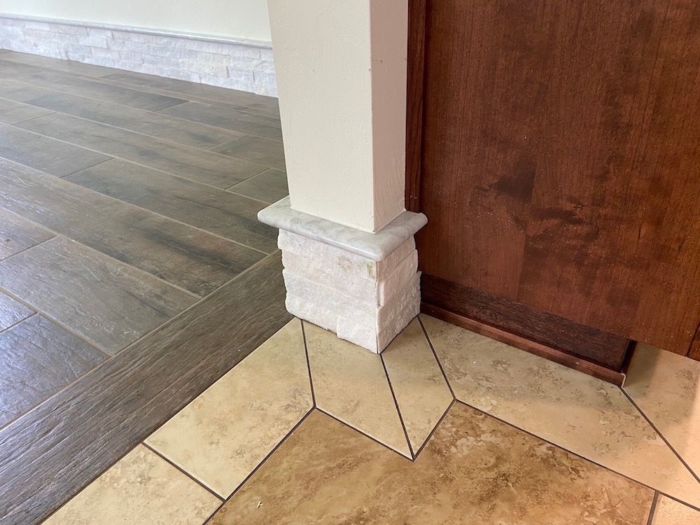 A square white column with stone veneer base sits on a two-toned tile floor next to a dark wood cabinet.