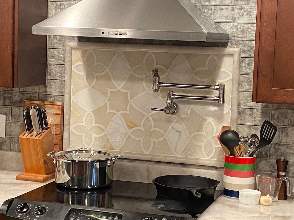 Kitchen with a stovetop, pot, pan, and tile backsplash featuring a pot filler, range hood, and wooden cabinets.