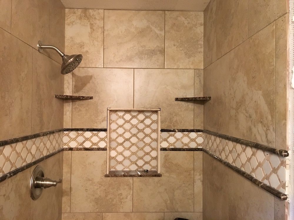 Shower stall with beige tile, decorative mosaic tile trim, built-in shelves, and a shower head.