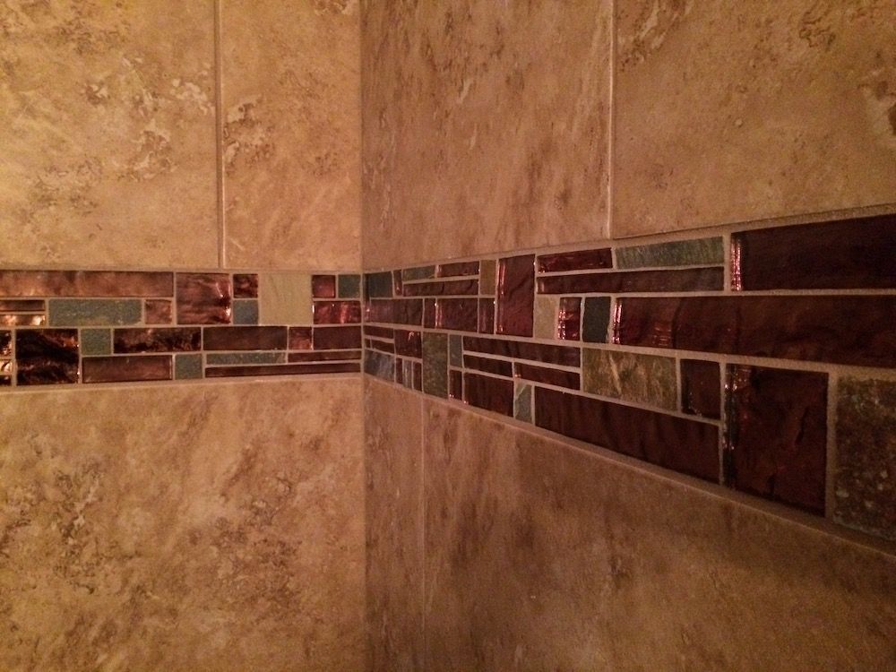 Corner of a shower with brown and tan tiles. Decorative mosaic tile border in shades of brown and green.