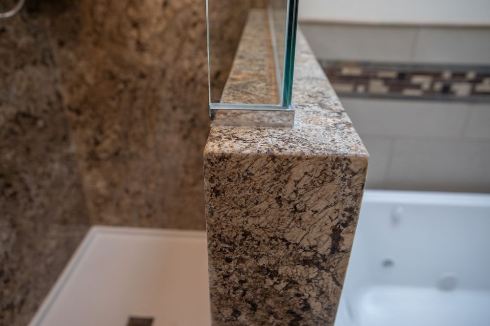 Close-up of a granite wall with a glass shower panel. Beige and brown tones are dominant. A white bathtub is in the background.