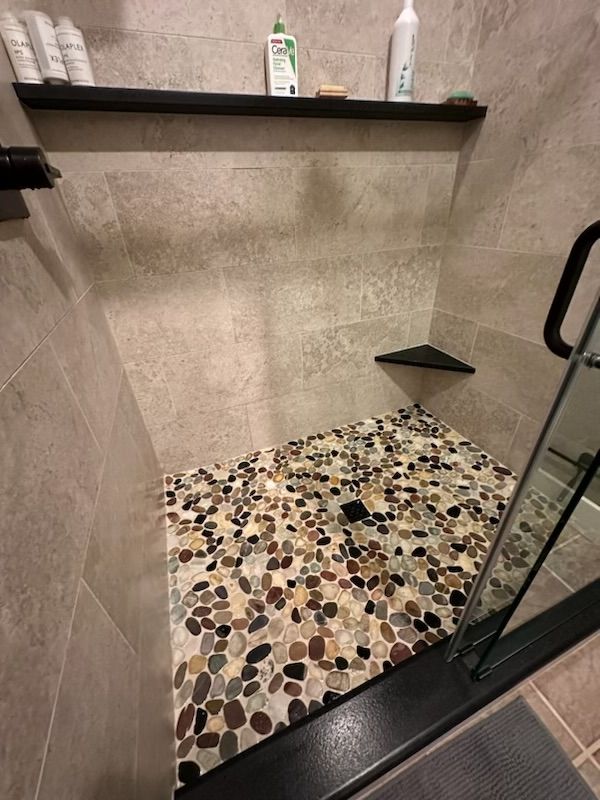 Shower with pebble floor, stone walls, black shelves holding toiletries, and glass door.