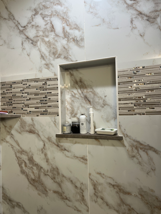 Marble-tiled shower with built-in shelf. Contains toiletries and a mosaic accent.