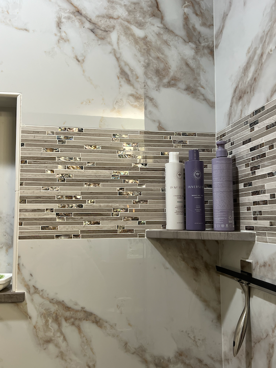 Shower with marble-look walls and mosaic tile accents. A shelf holds shampoo bottles.