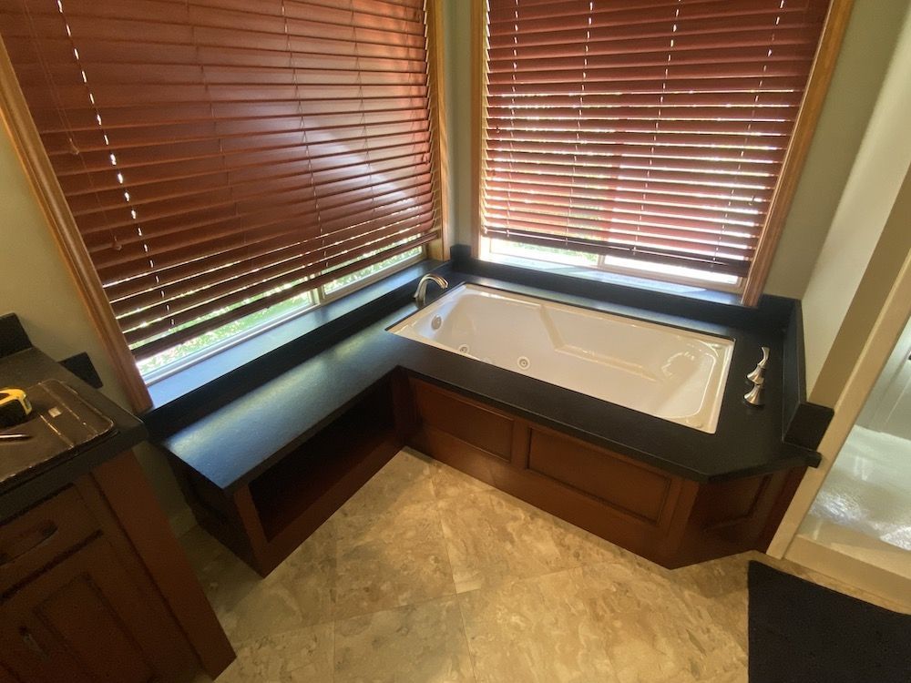 Bathroom corner with built-in jacuzzi tub, dark wood cabinets, black countertops, wood blinds.