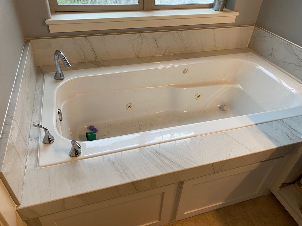 White bathtub with water jets, set in marble tile surround, with chrome fixtures.