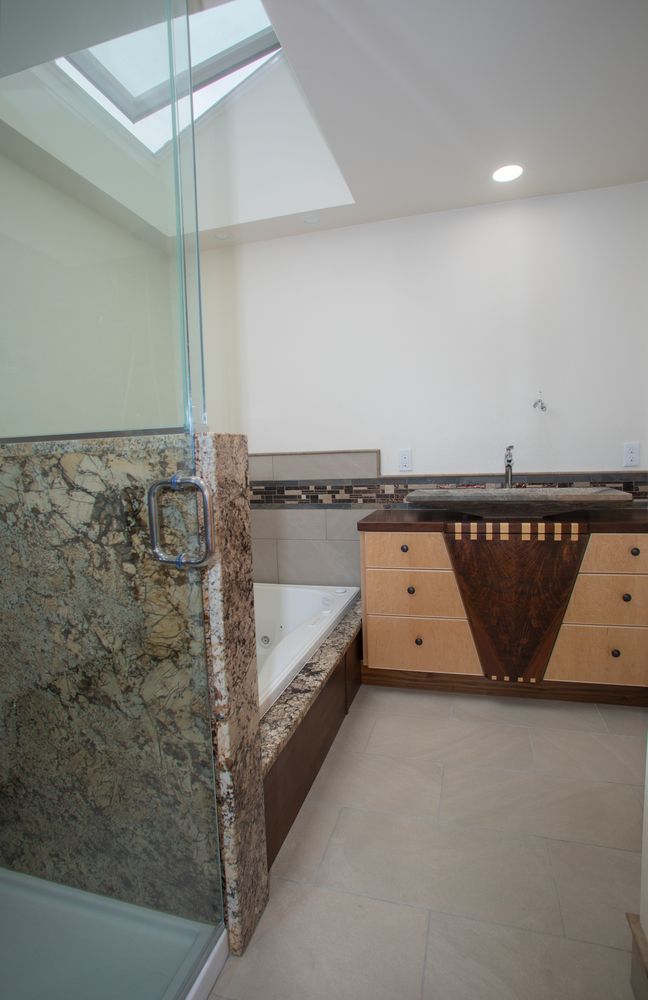 Bathroom with granite-walled shower, jacuzzi tub, and vanity with dark countertop and light wood cabinets, under a skylight.