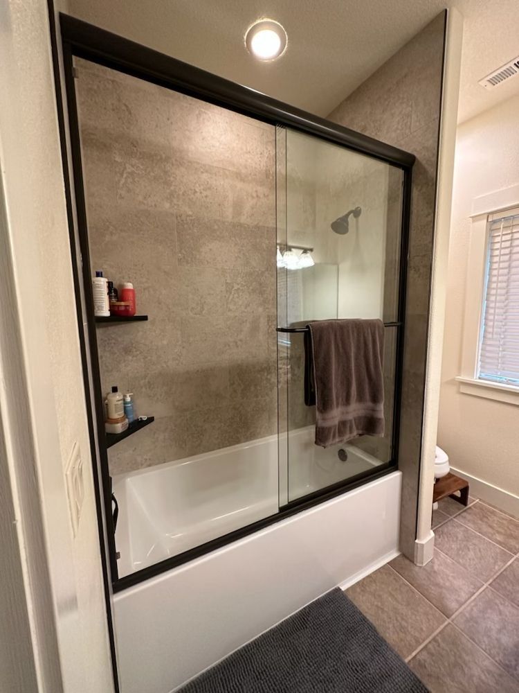 Bathroom with white tub, glass shower door, towel, brown and gray tile walls, and a window.
