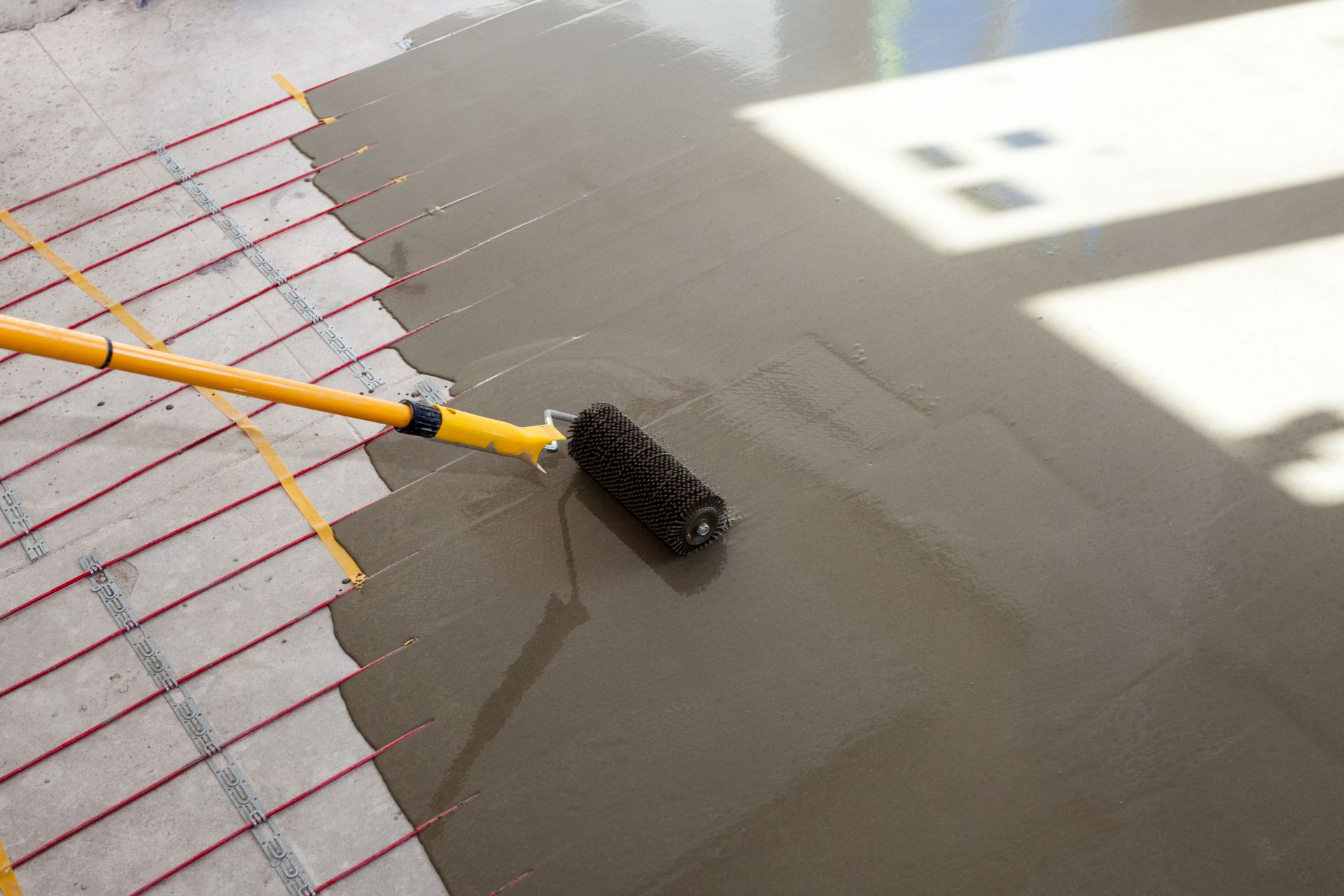 A spiked roller on a long yellow handle is being used to smooth wet, self-leveling compound over an interior floor.
