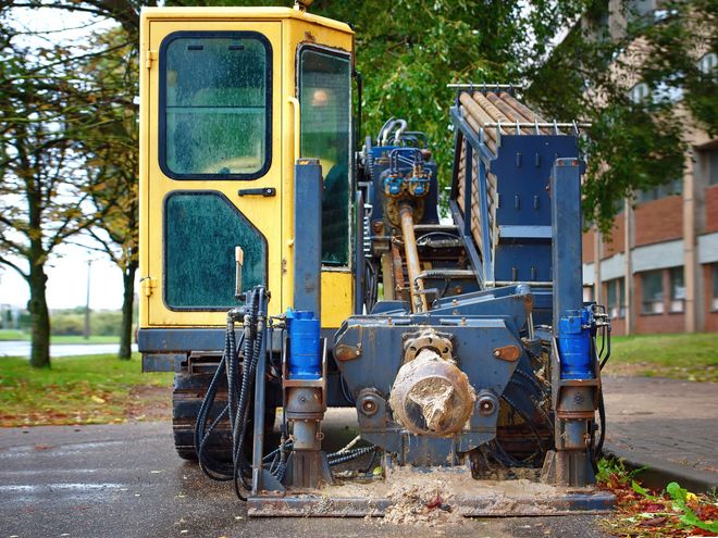 Yellow directional drilling rig parked on pavement with trees and a brick building in the background.
