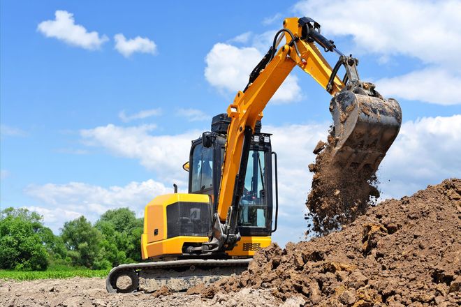 A yellow excavator sits in a field under a blue sky, using its bucket to move a pile of dirt.