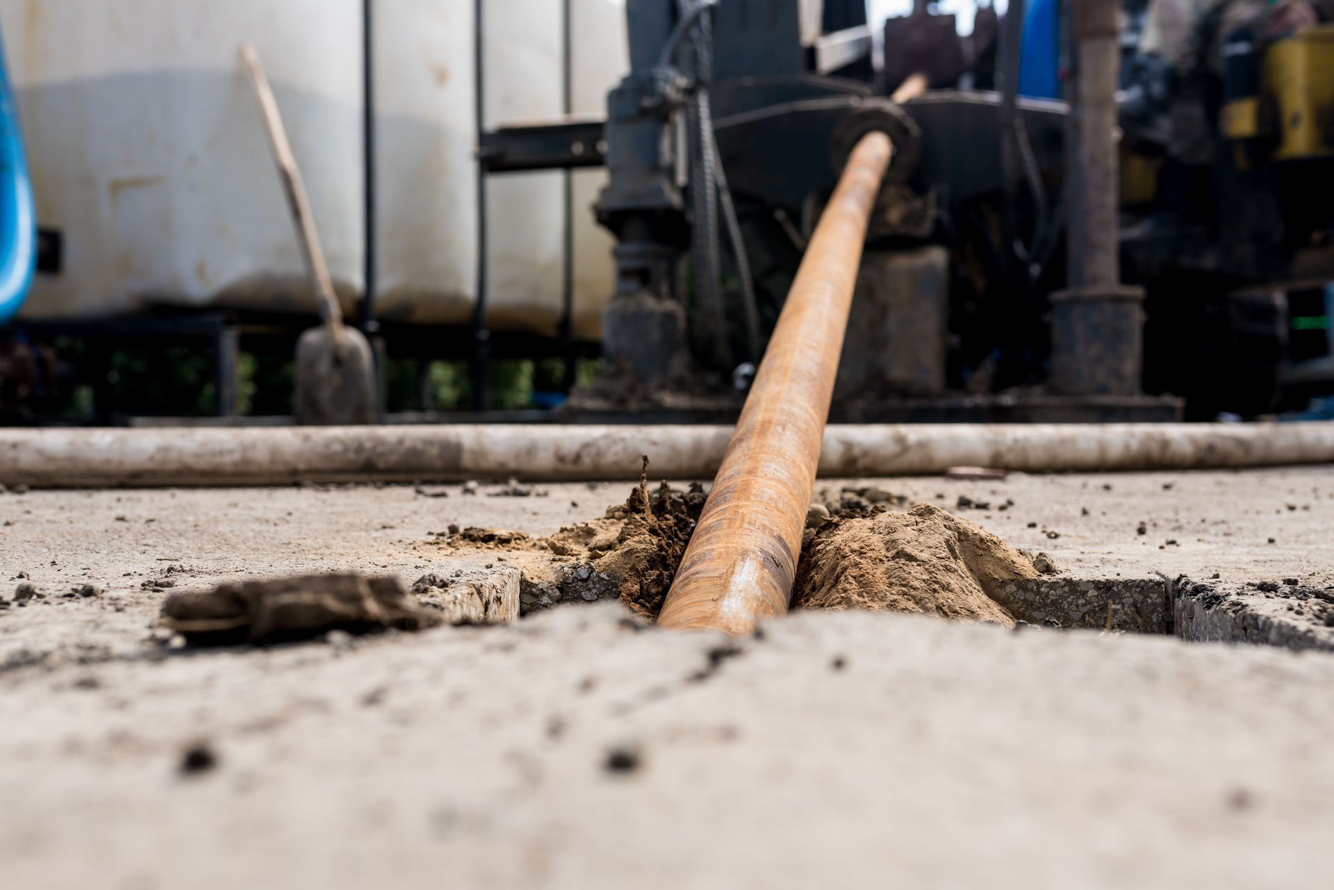 A metal drill pipe extending into a hole in concrete at a job site with industrial equipment in the background.