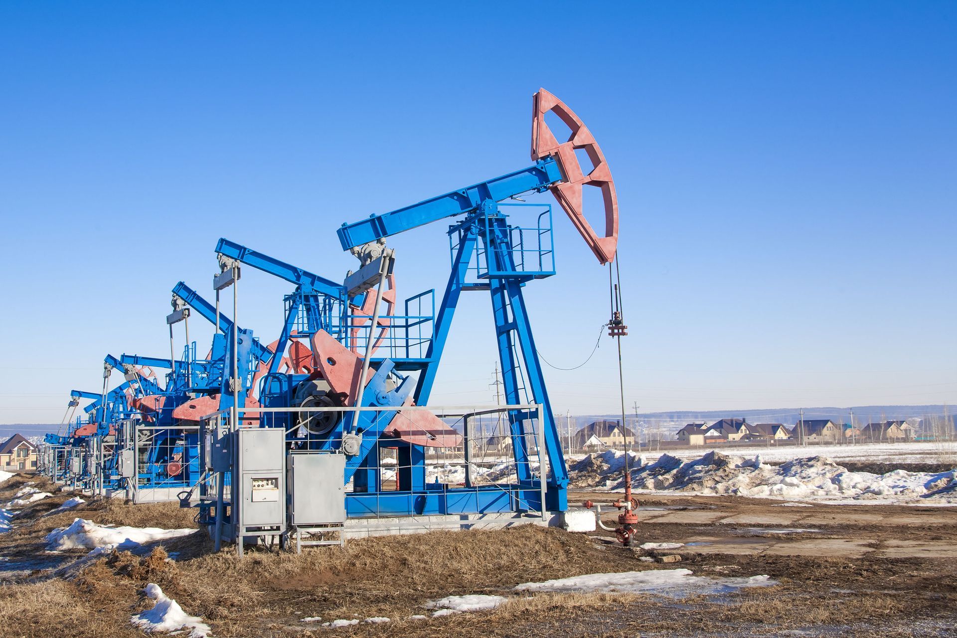 A row of blue oil pump jacks operating in a snowy, rural landscape under a clear blue sky.