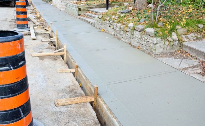A newly poured concrete sidewalk under construction, bordered by wooden forms and orange traffic barrels.