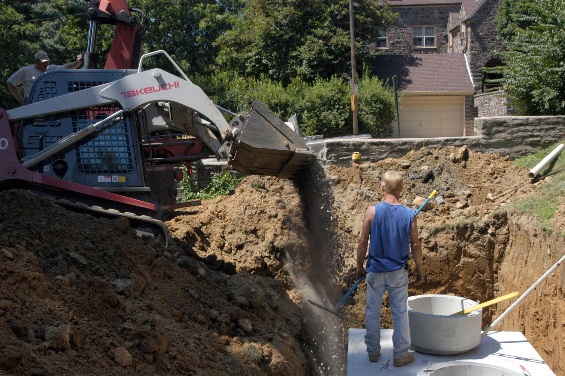 A construction worker stands in a deep trench while a skid-steer loader dumps dirt near a concrete drainage structure.