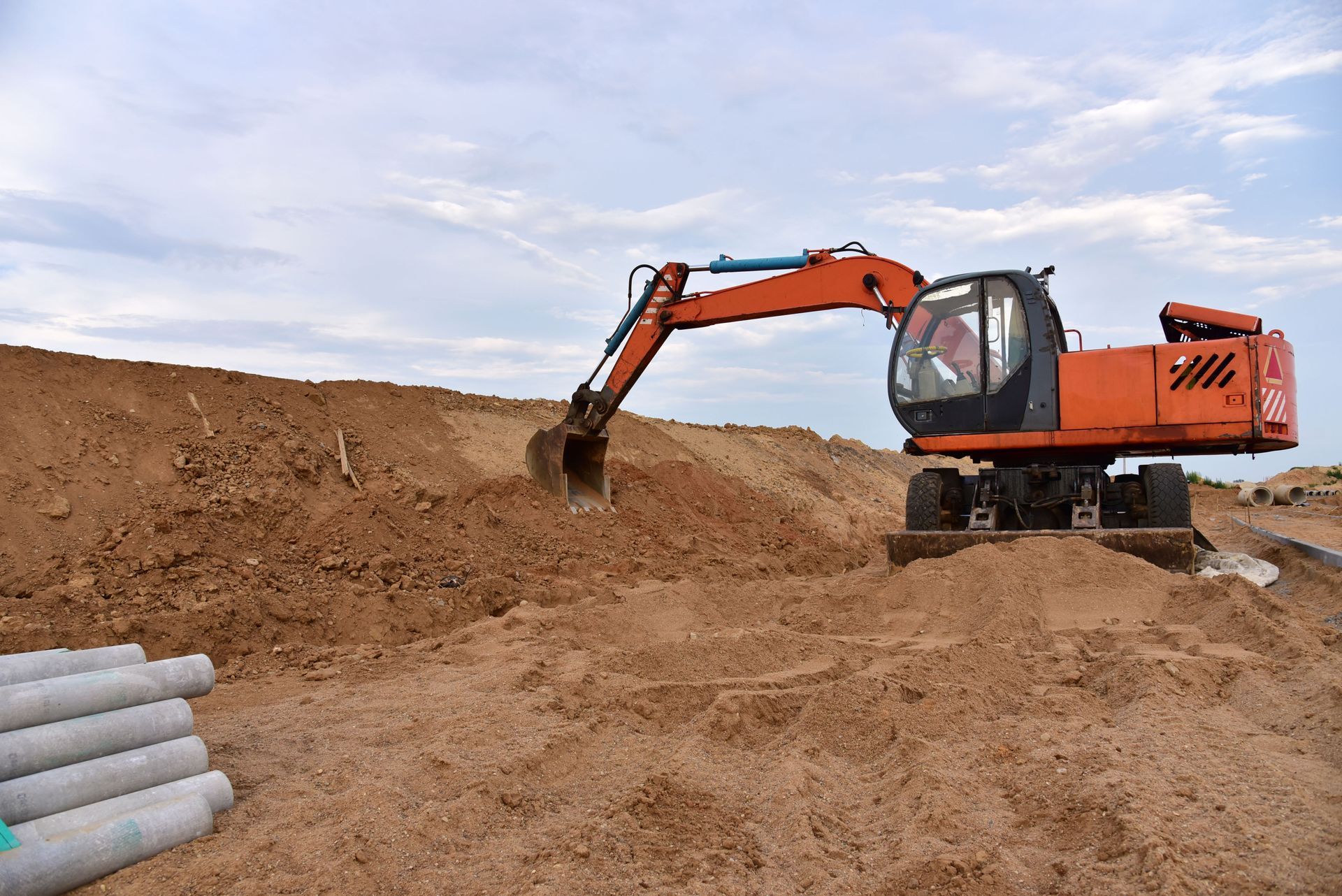 An orange excavator sits on a dirt mound next to a pile of concrete pipes at a construction site under a cloudy sky.