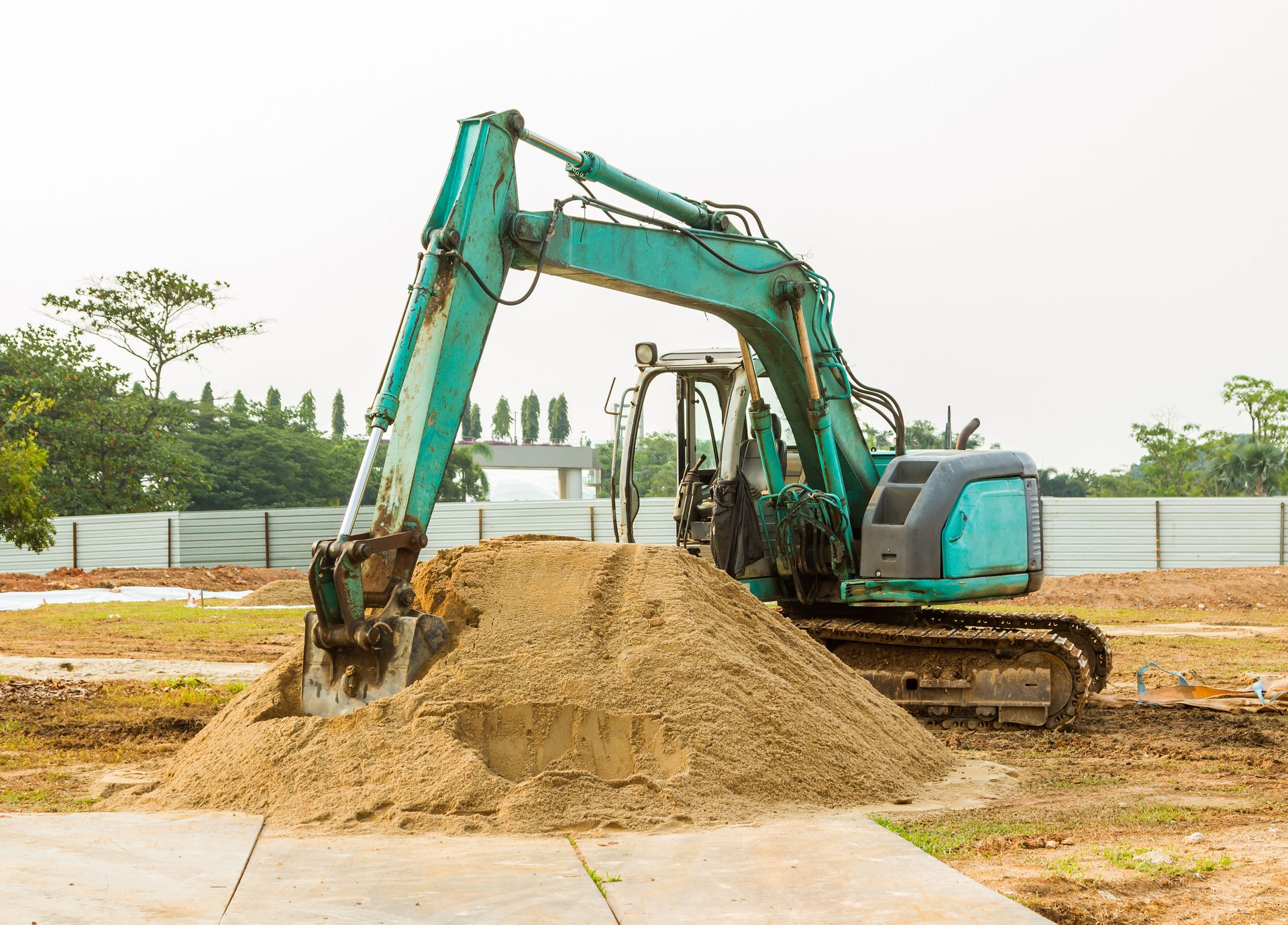 A teal excavator sits on a dirt lot, digging into a pile of sand under a pale, overcast sky.