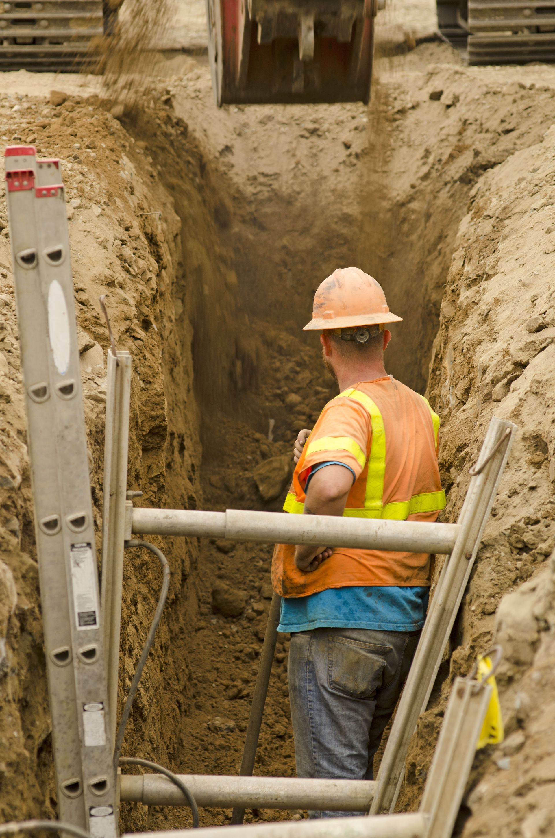 Worker in orange vest and hard hat standing in a deep trench beside excavation supports.
