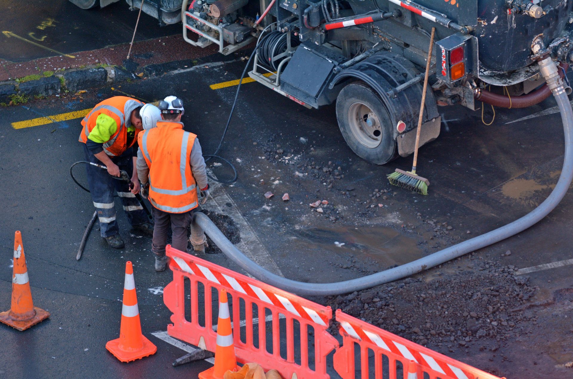 Two workers in high-visibility orange vests use a vacuum truck to clear debris from a road excavation behind safety cones.
